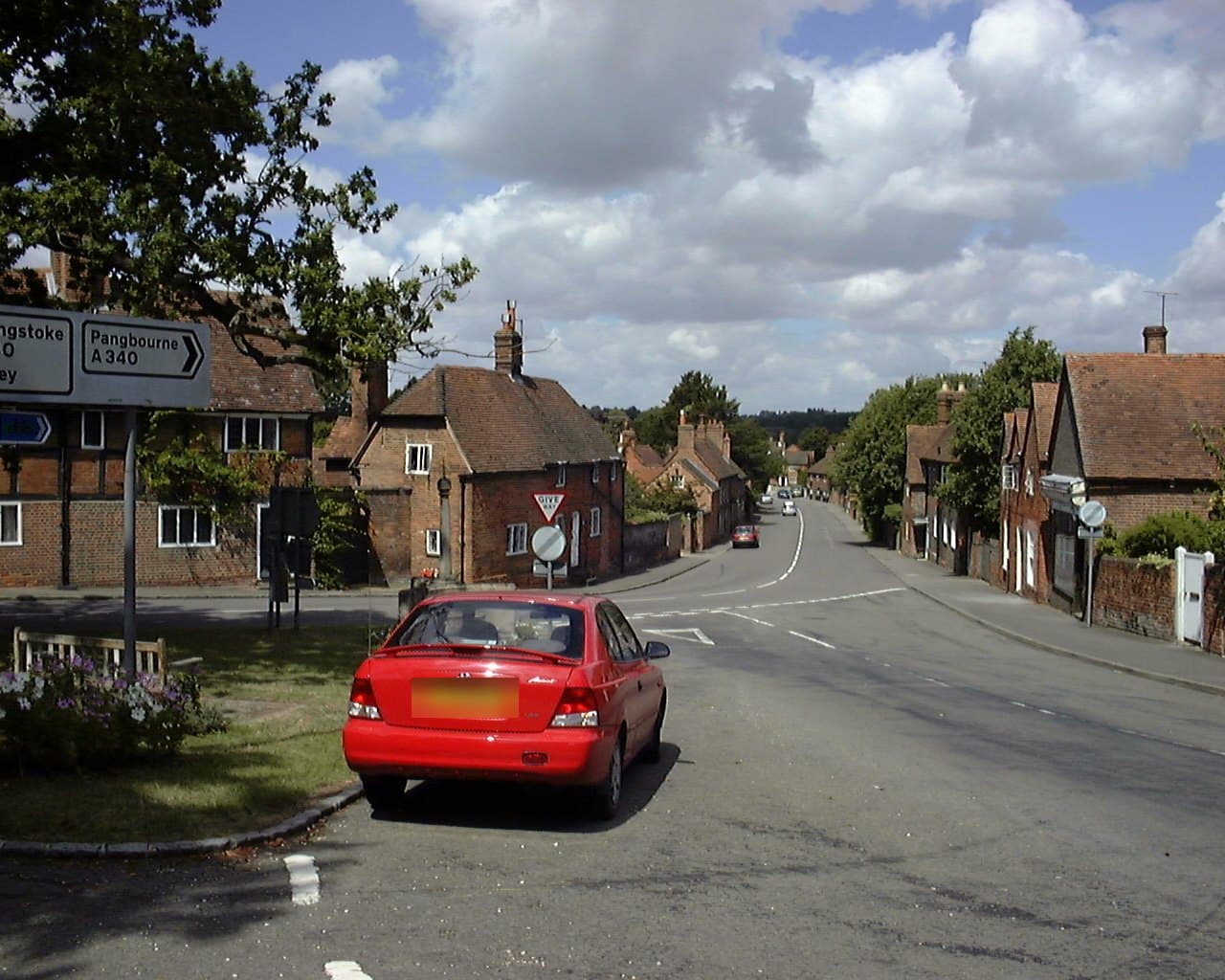 Aldermaston's village street running away downhill, lined with old red brick and timber cottages, with a signpost for the A340 and a tree on the green in the foreground.