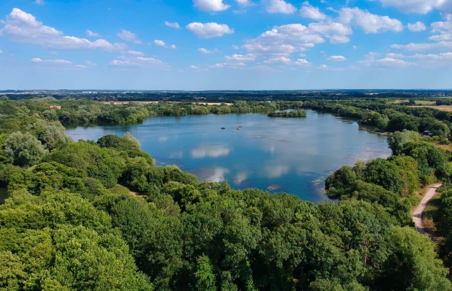 A large lake ringed by dense summer woodland, seen from the air, with white clouds reflected in the still water and open country beyond.