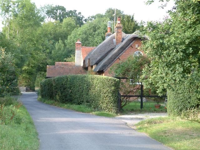 A thatched brick cottage with tall chimneys behind a clipped hedge, at a bend in a narrow lane under trees.